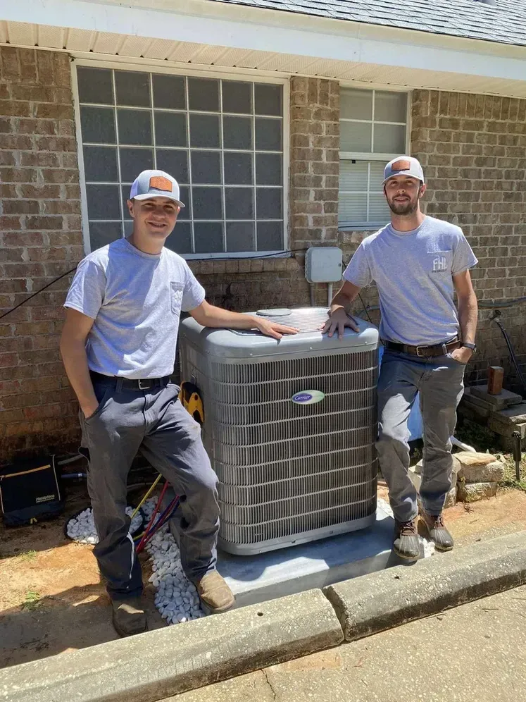 Two HVAC technicians posing beside a new outdoor air conditioner unit outside a brick house