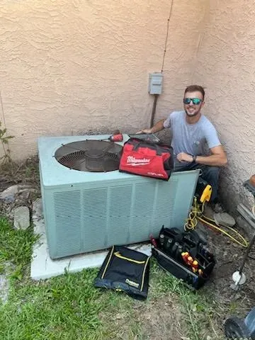 Man kneels beside outdoor HVAC unit, holding tools and a red bag against a stucco wall.