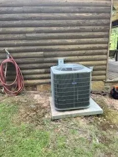 Outdoor air conditioner unit on a concrete pad beside a log cabin wall, with a red hose coiled nearby.