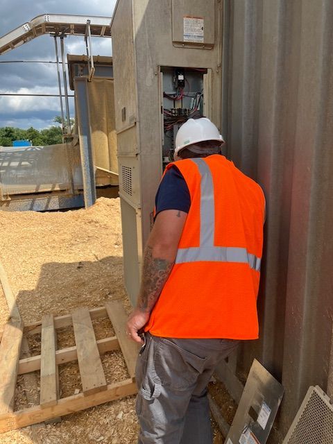 Worker in orange safety vest and white hard hat inspecting equipment beside industrial tanks.