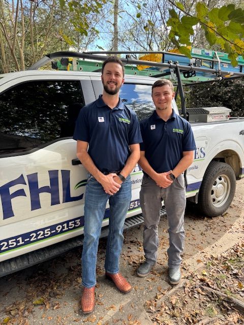 Two men in navy shirts standing beside a white FHL service truck with ladders on top outdoors