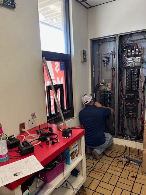 Technician kneels beside an open electrical panel in a utility room, with tools on a red workbench nearby.