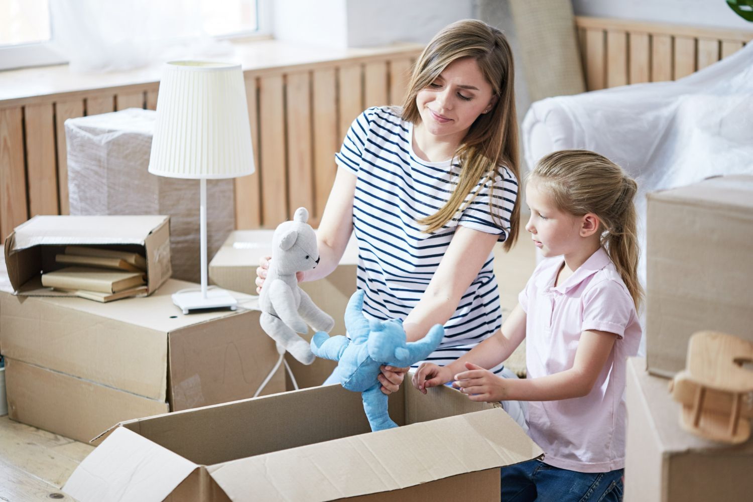 Two children unpack stuffed animals from a cardboard box in a bedroom