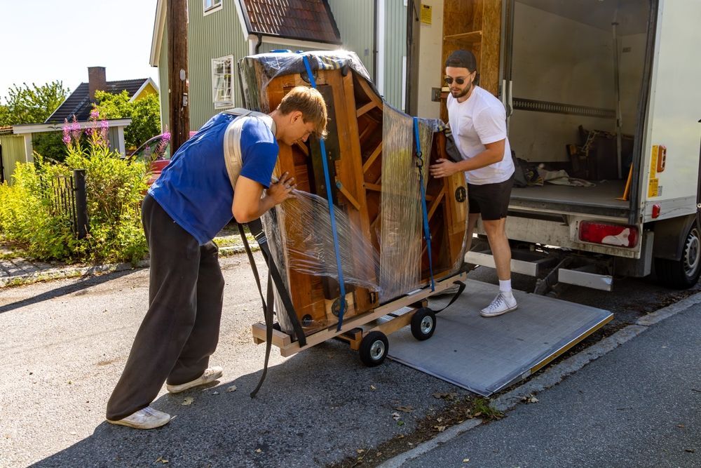 Two workers unloading a wrapped upright piano from a truck using a dolly and ramp