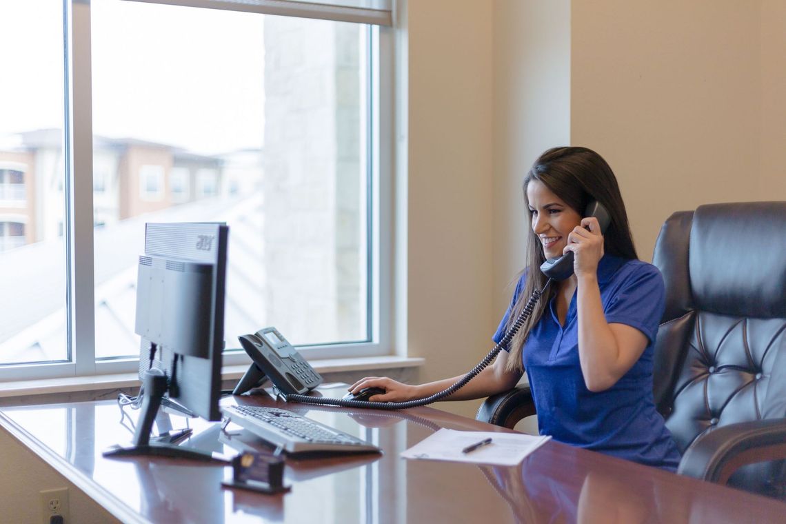 Woman in blue blouse talking on the phone at a desk with a computer in a bright office