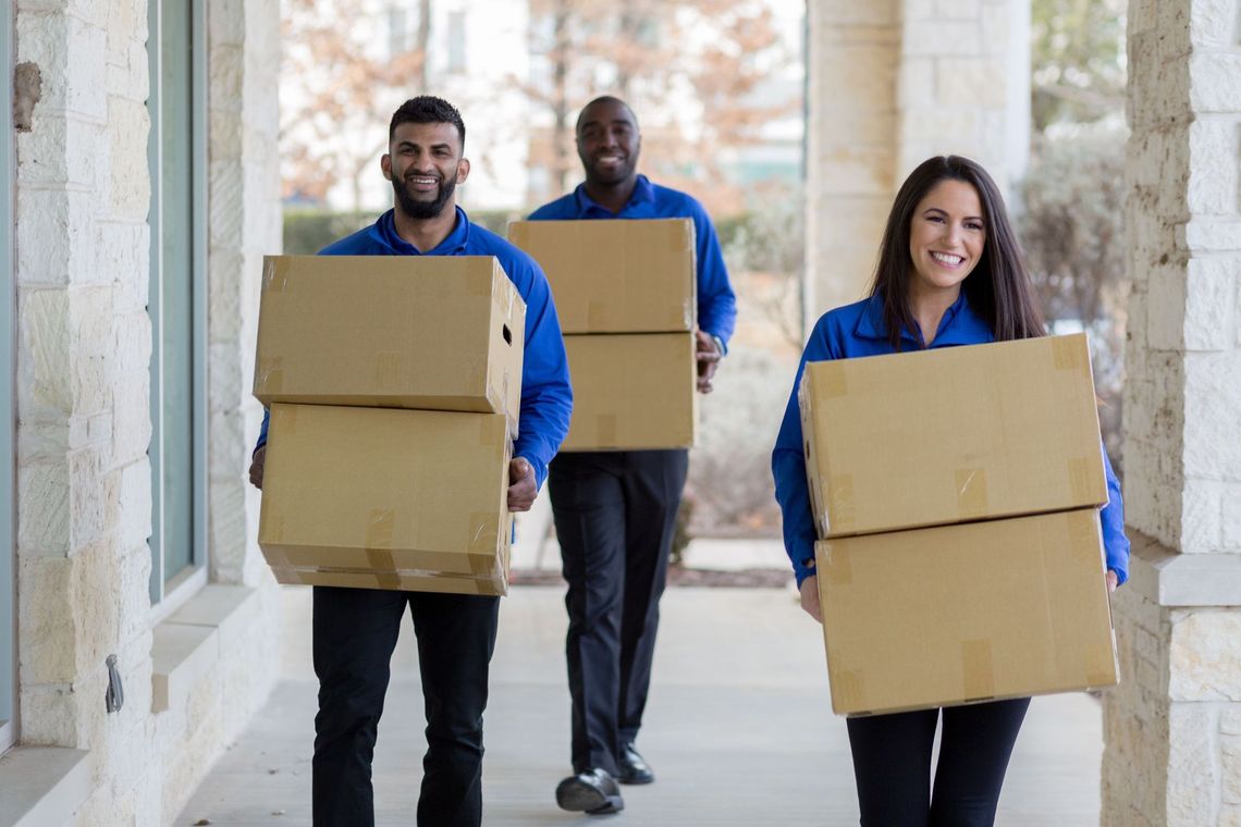 Three people carrying cardboard boxes down a bright hallway, smiling as they move.