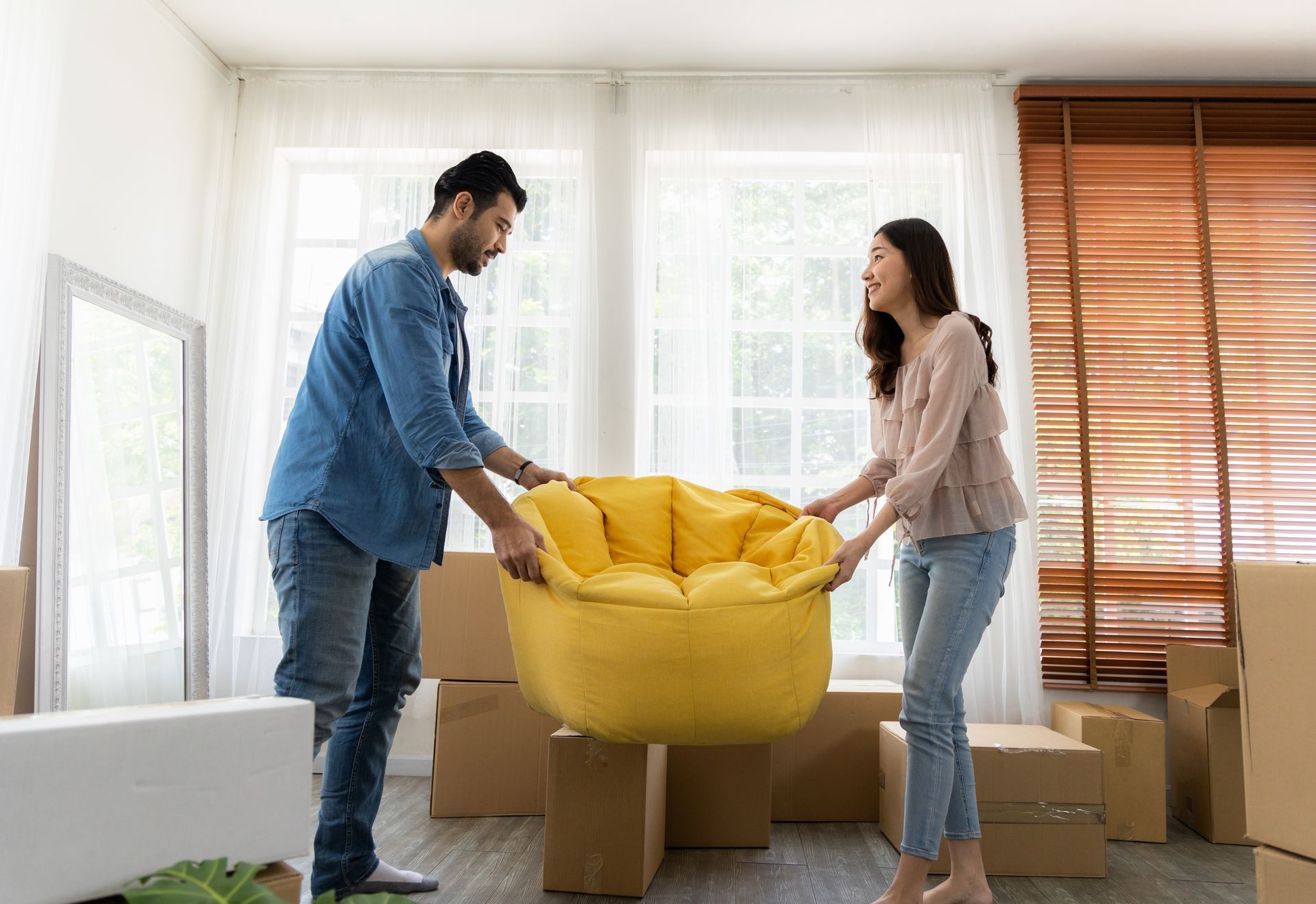 Couple moving, carrying a yellow armchair among cardboard boxes in a room with a window and blinds.