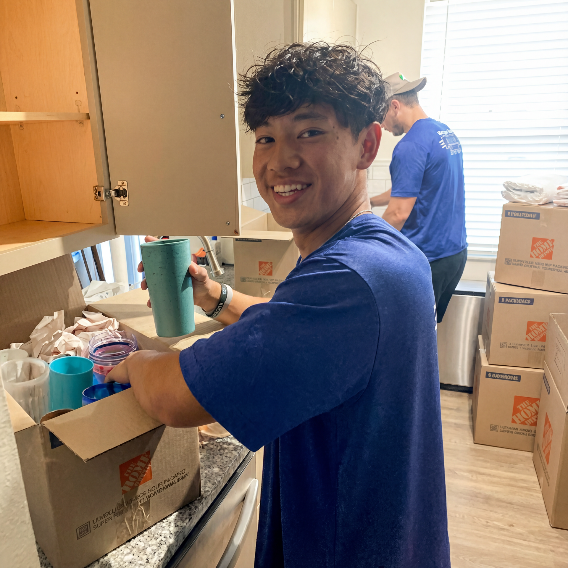 Smiling person in a blue shirt packing boxes in a sunlit kitchen.