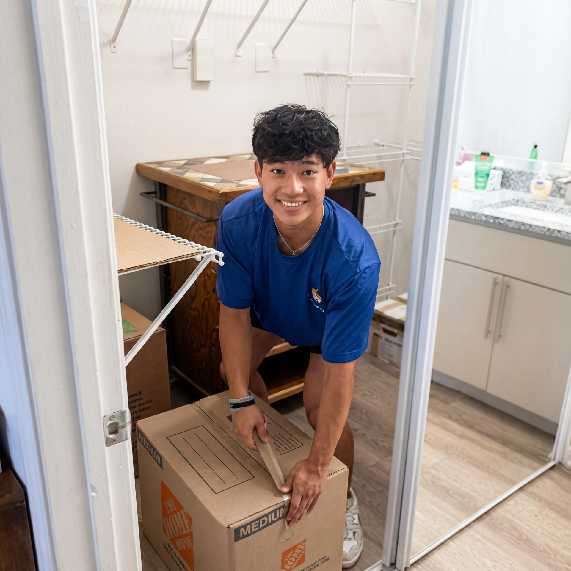 Person in a blue shirt carrying a cardboard box in a closet-like storage room.