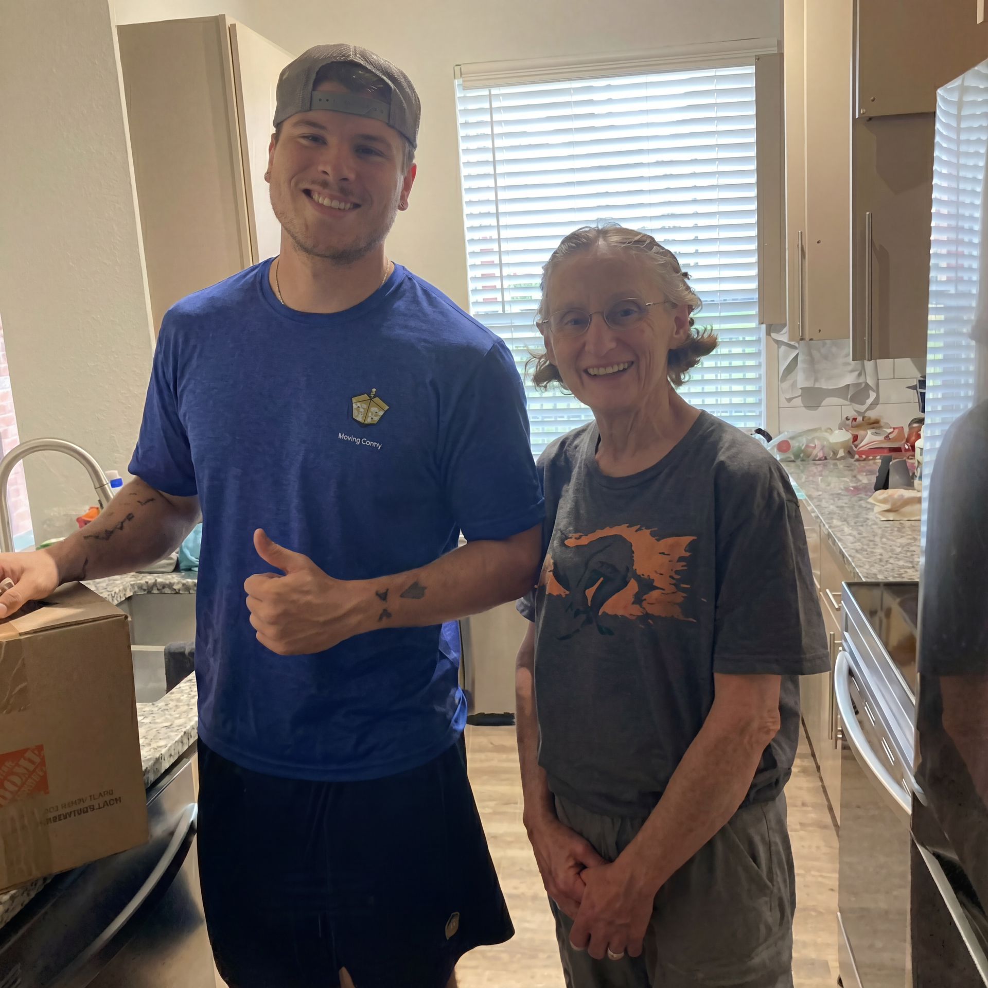 Two people smiling in a kitchen, one holding a cardboard box near a countertop.