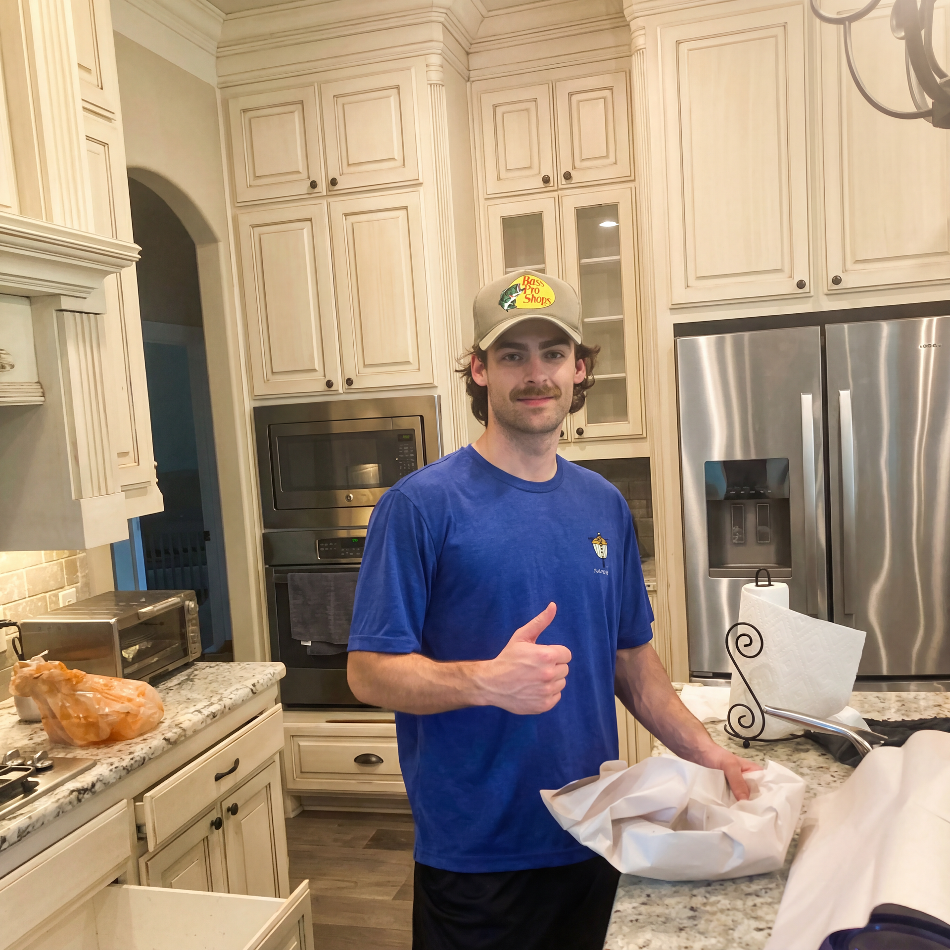 Man in a blue shirt and cap giving a thumbs-up in a bright kitchen, standing by a counter with food.