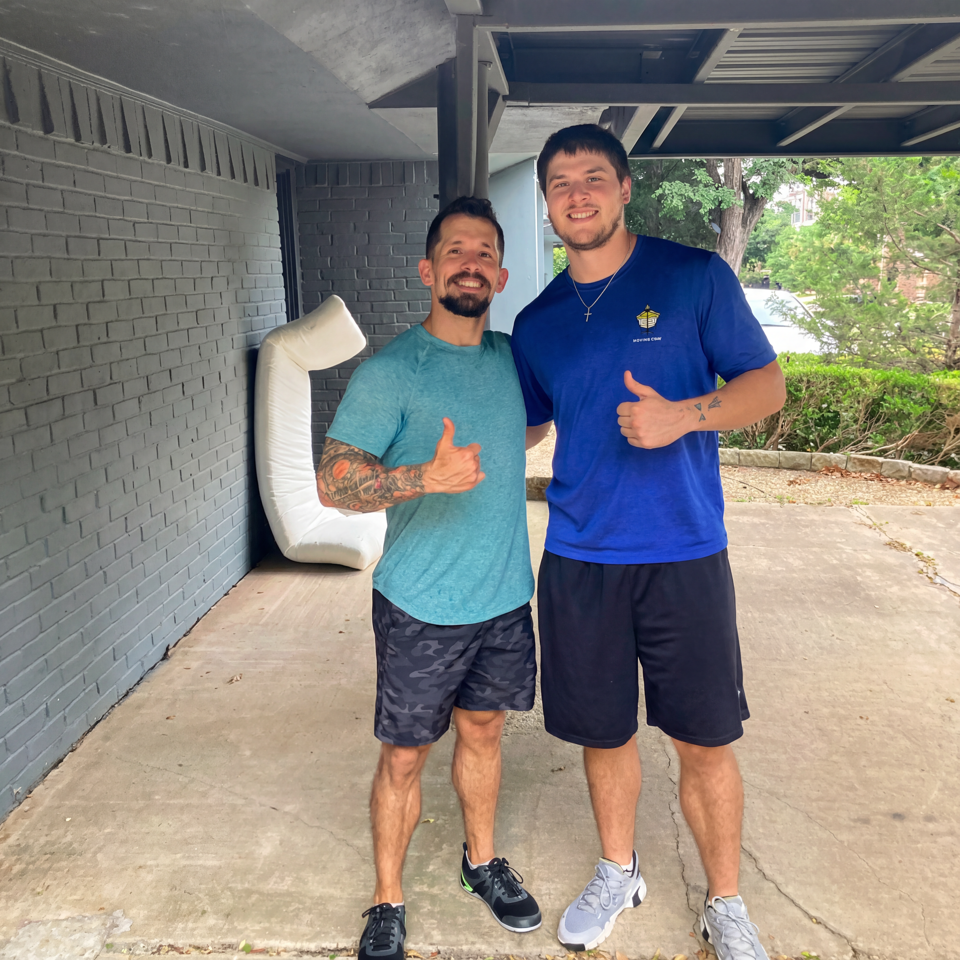Two men smiling and standing under a covered walkway, one giving a thumbs-up.