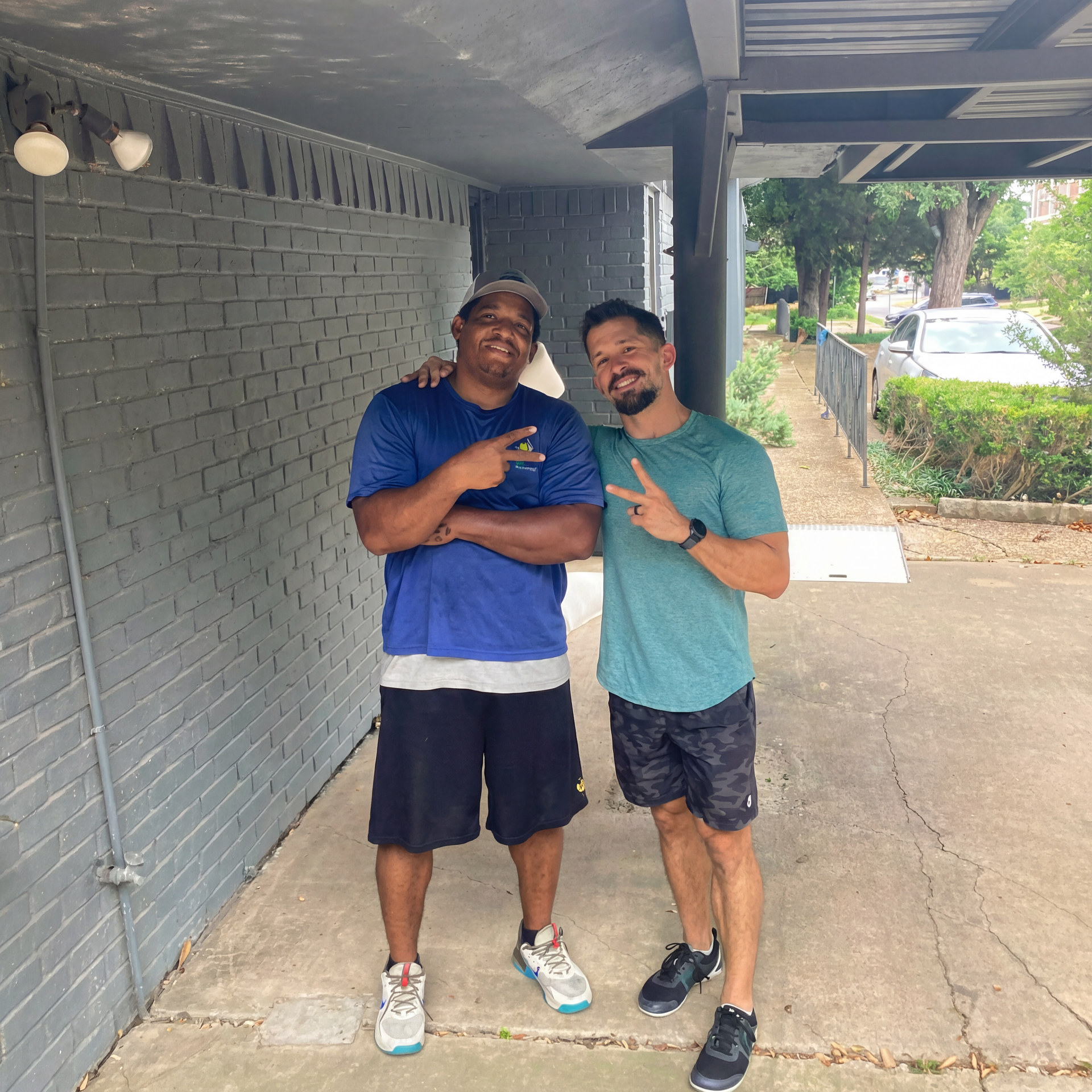Two people smiling and posing under a covered walkway, with one brick wall and a sidewalk beside them.