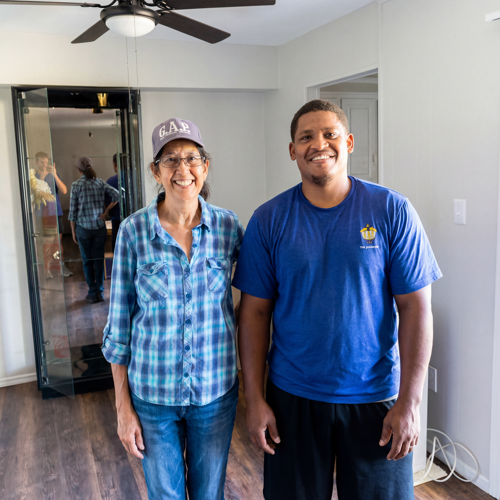 Two people smiling and standing indoors in a room with a ceiling fan and mirror.