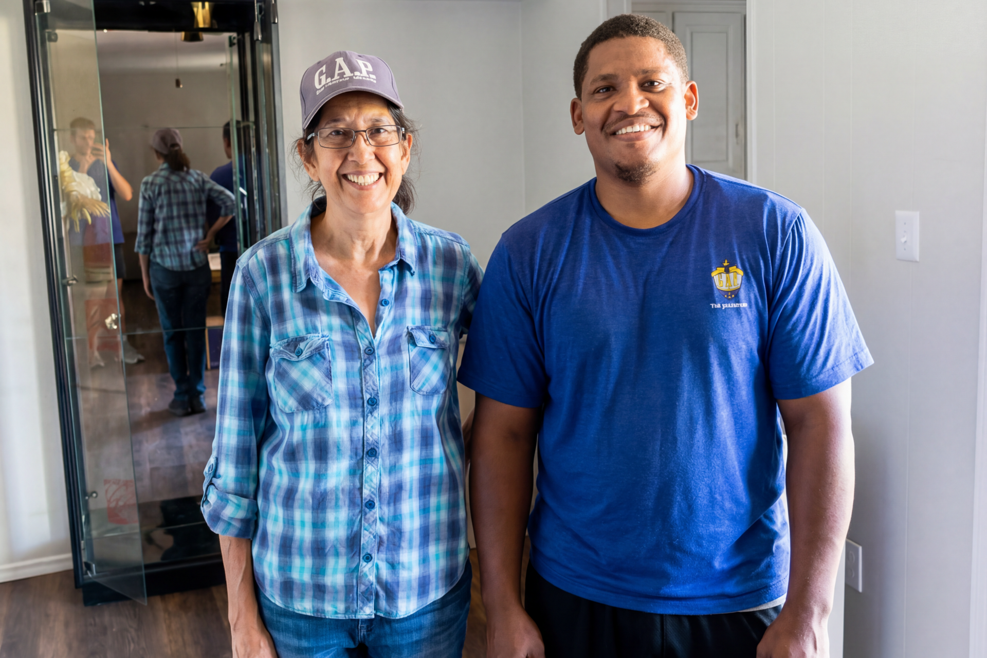 Two smiling people standing indoors, one in a blue plaid shirt and cap, the other in a blue T-shirt.