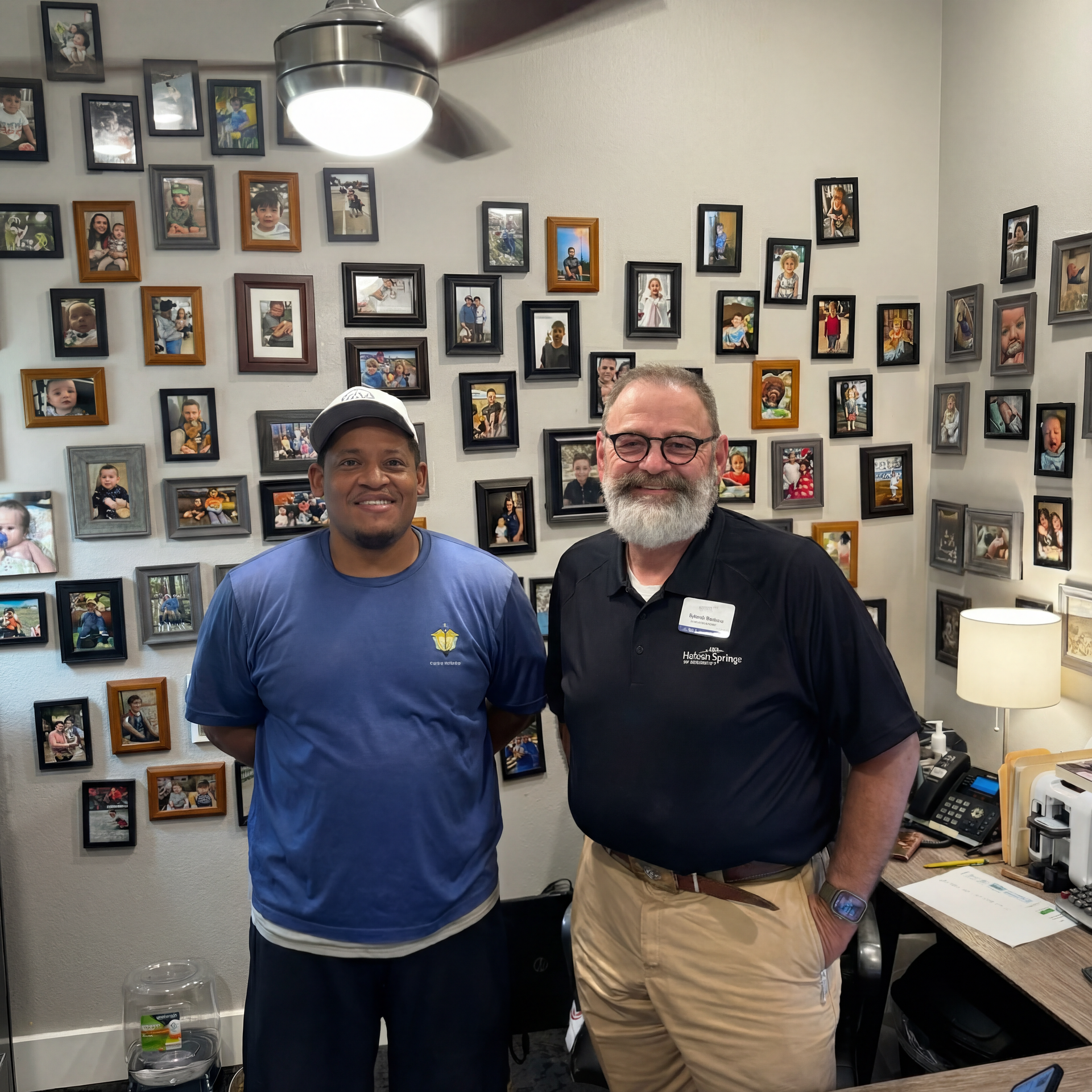 Two smiling people standing in a room with a wall covered in many framed photos