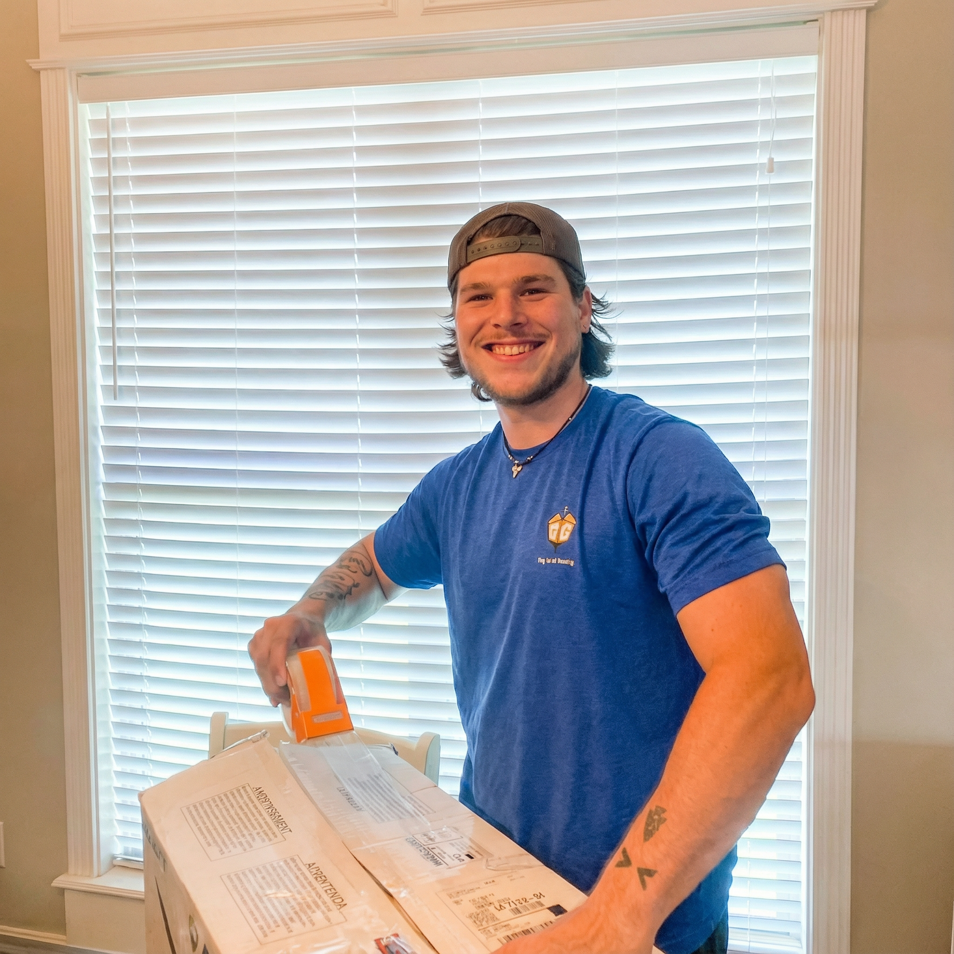 Smiling man in a blue shirt painting a white wall inside a room with blinds covering the window