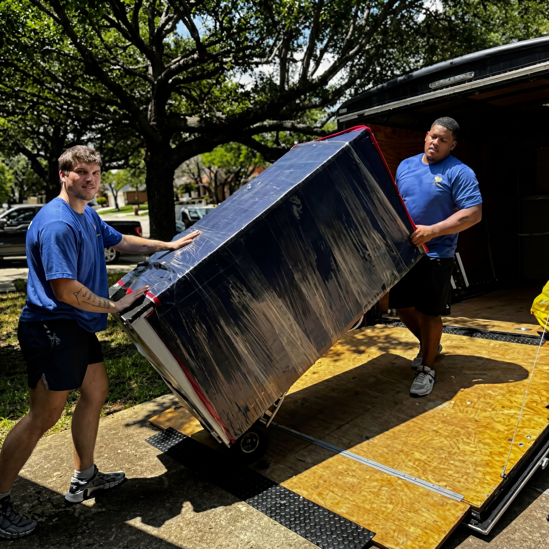 Two men in blue shirts loading a large metal box onto a truck ramp outdoors.