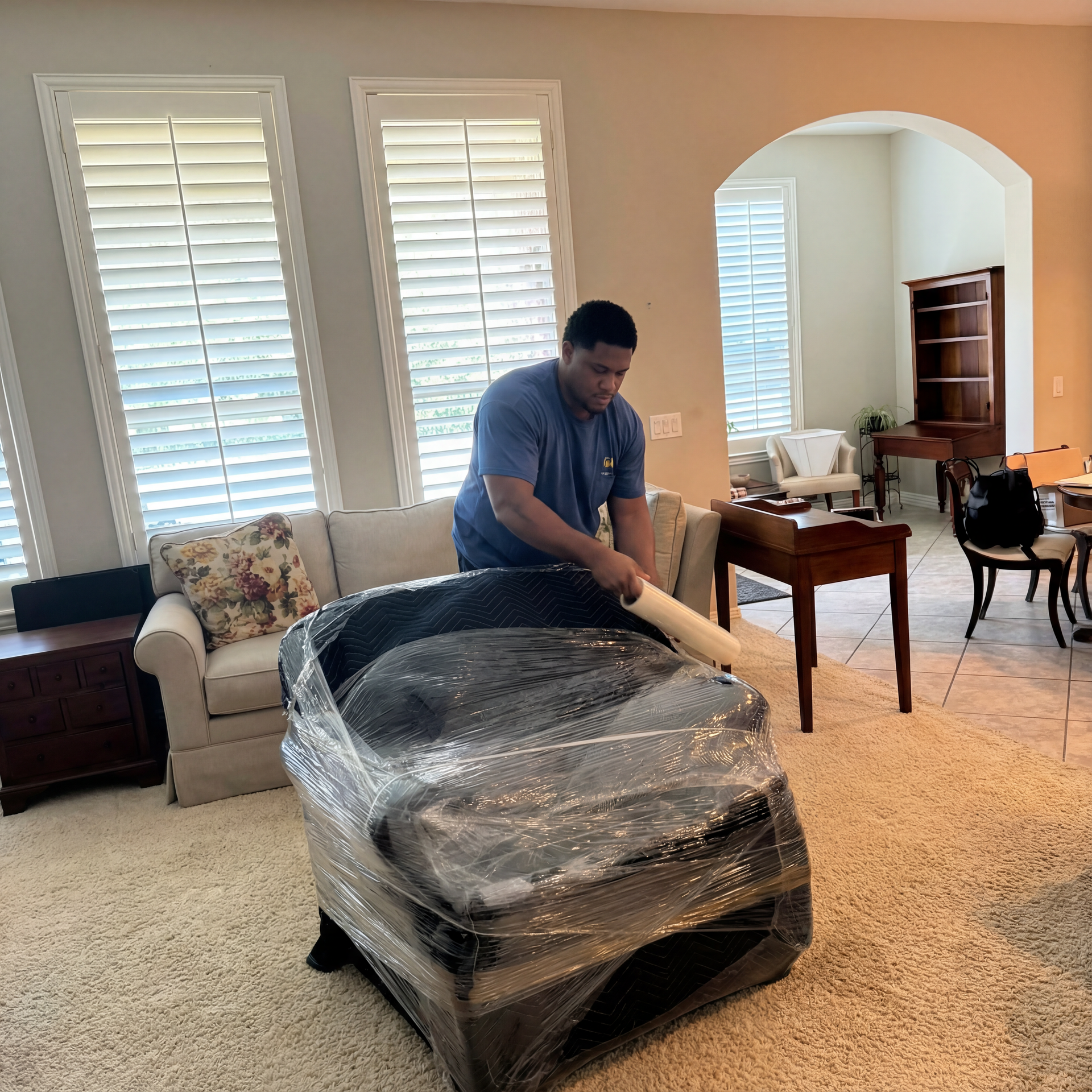 Person unwrapping a black armchair in a bright living room with beige carpet and white shutters