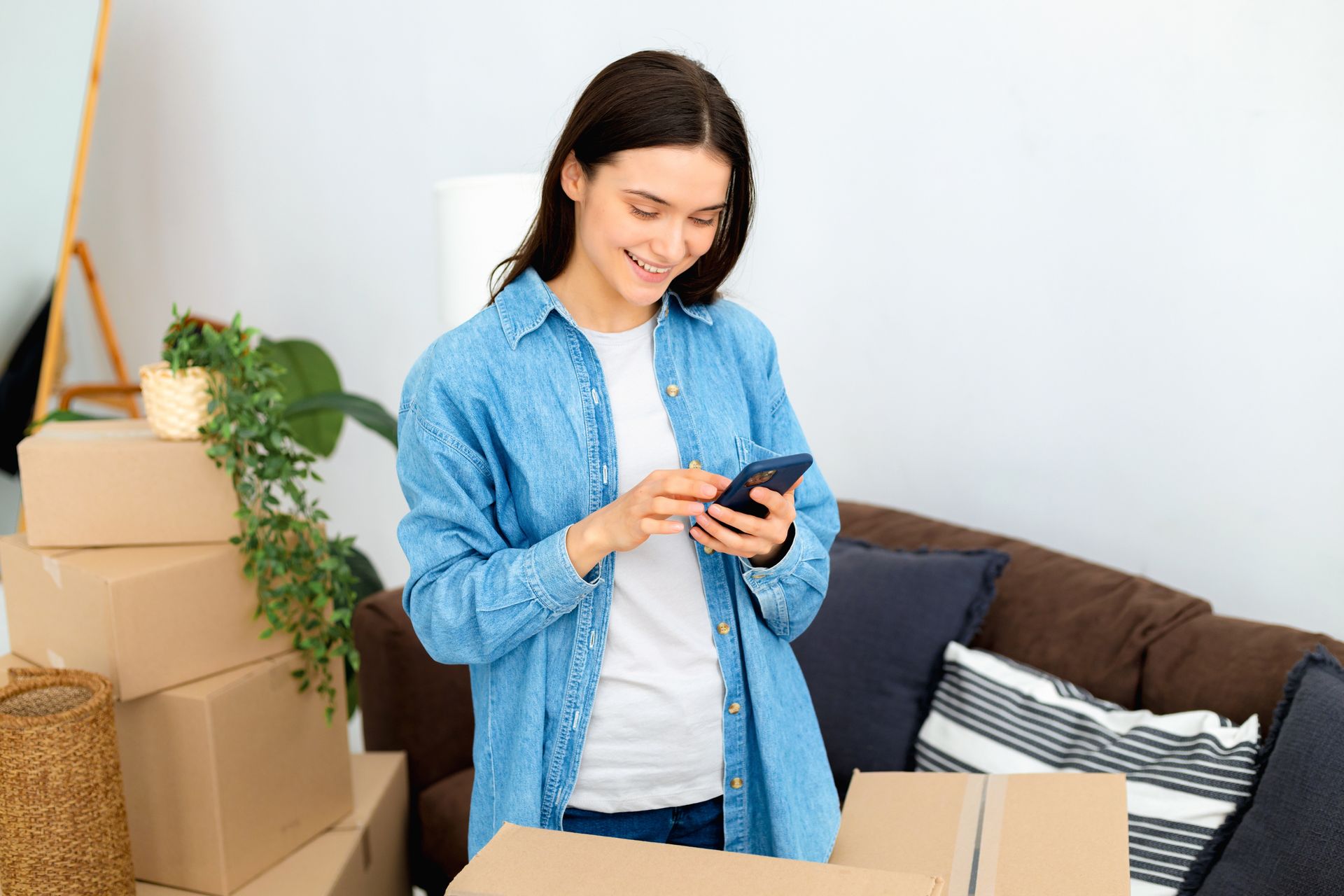 Woman in a denim shirt uses a phone, smiling. Boxes and a couch are in a room.