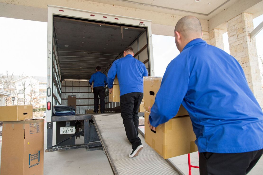 Movers in blue uniforms loading boxes into a truck via a ramp
