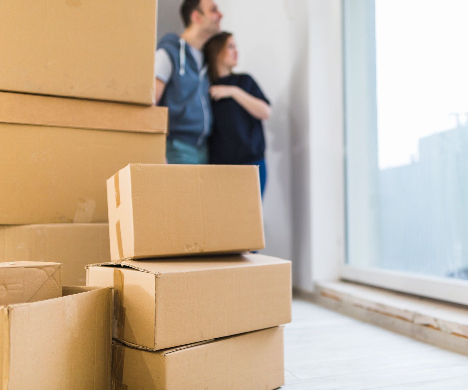 Moving boxes stacked in a bright room, with two blurred people standing near a large window.