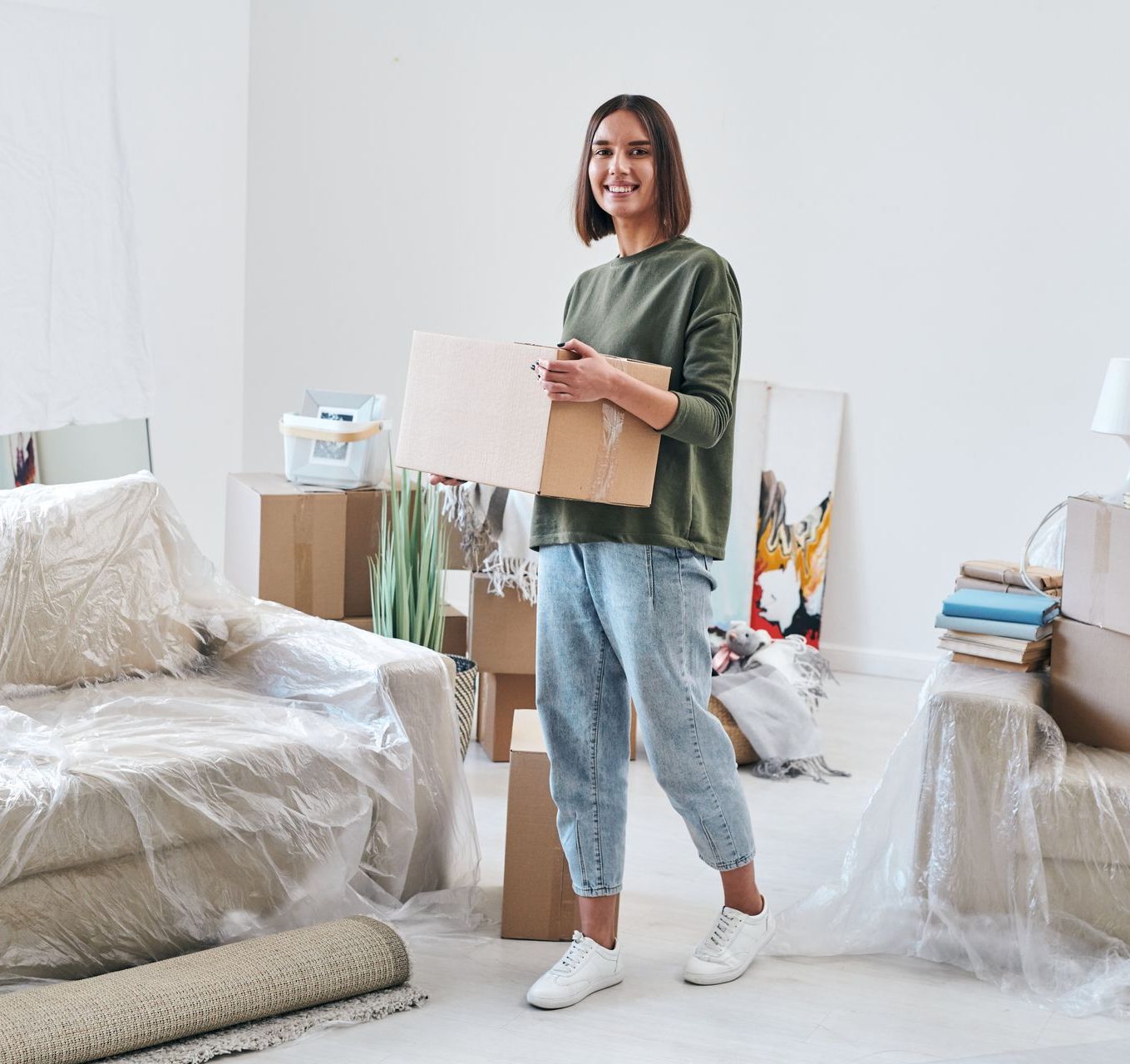 Woman holding a box in a bright, unpacked room with furniture covered in plastic