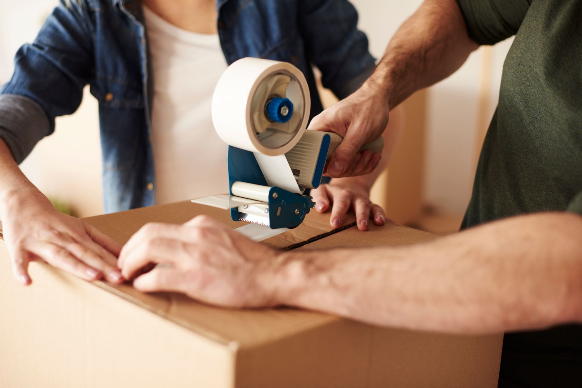 Hands taping a cardboard box with a packing tape dispenser at a table