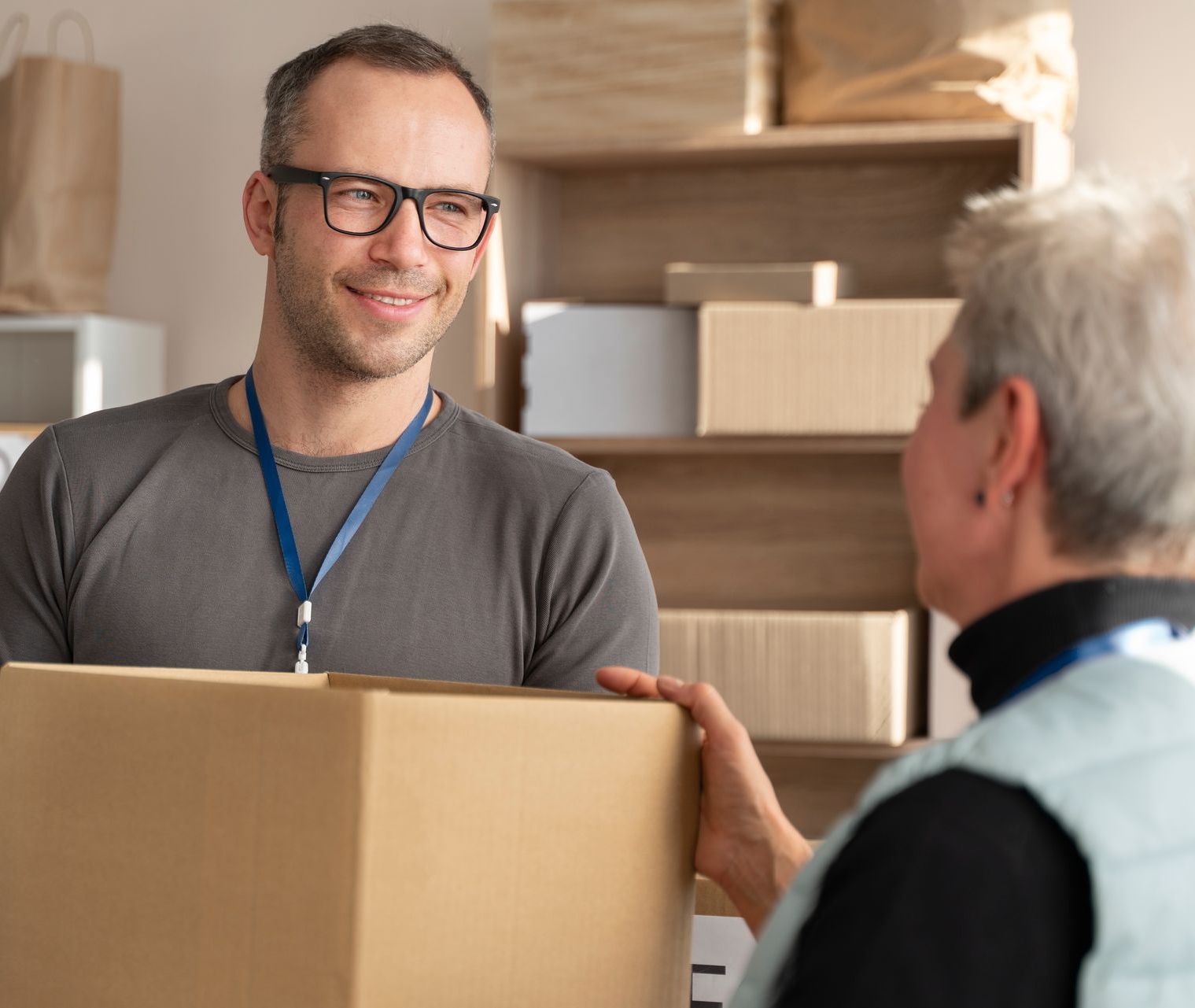 Two people exchanging a cardboard box in a storage room with stacked moving boxes.