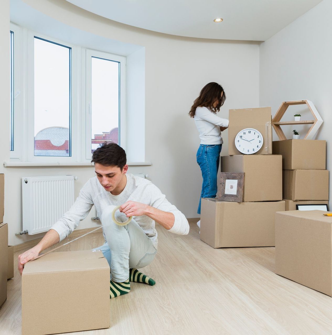 Two people unpacking cardboard boxes in a bright, empty room moving into a new home