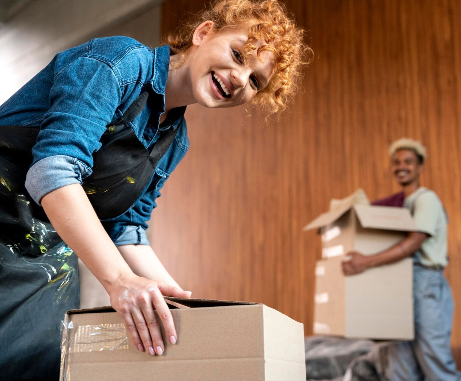 Two people packing boxes in a moving scene, one smiling in the foreground, wooden wall behind them