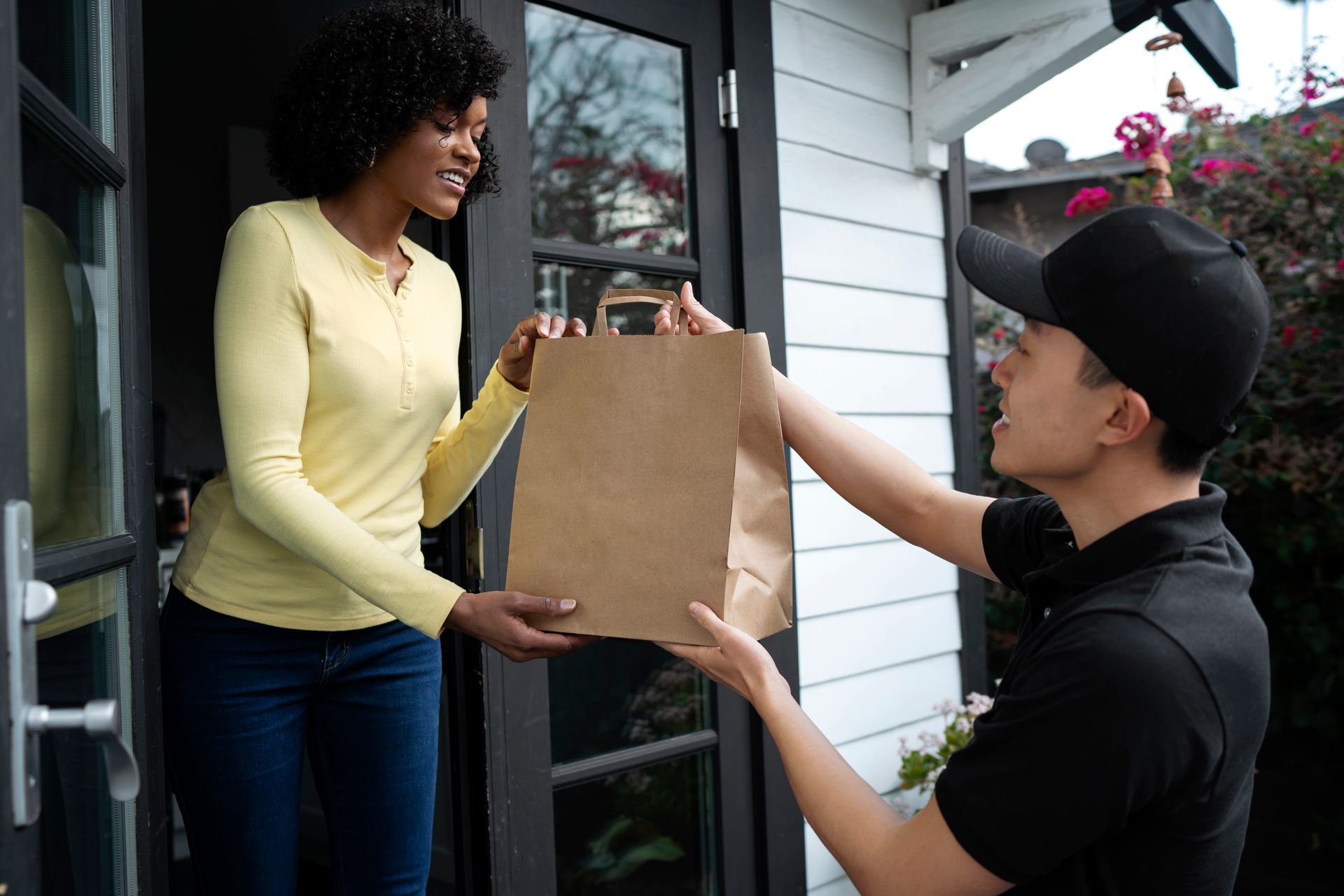 Person hands a paper grocery bag to a delivery worker at a front door.