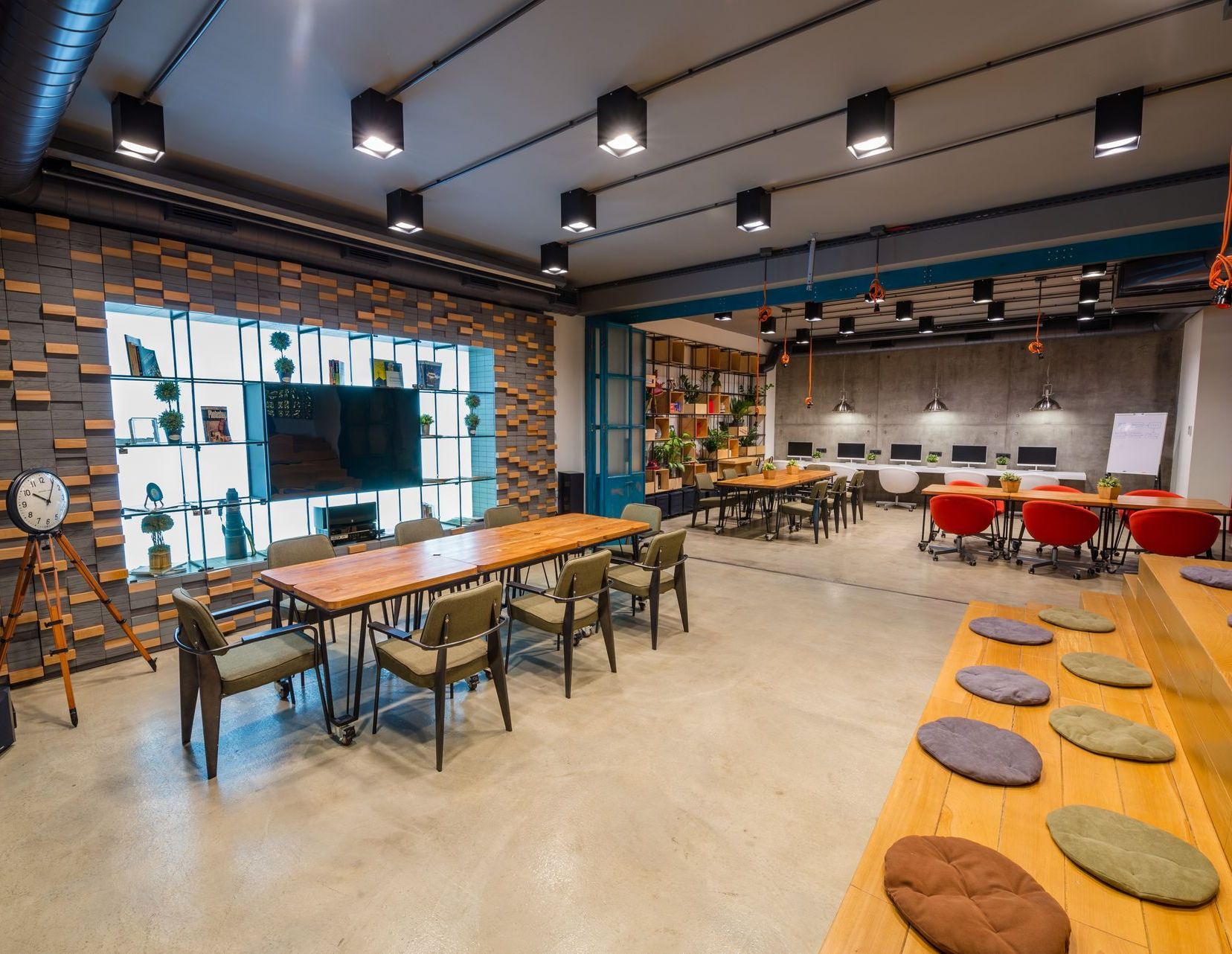 Modern open office lounge with long tables, lounge seating, wall TV, and red chairs in the back