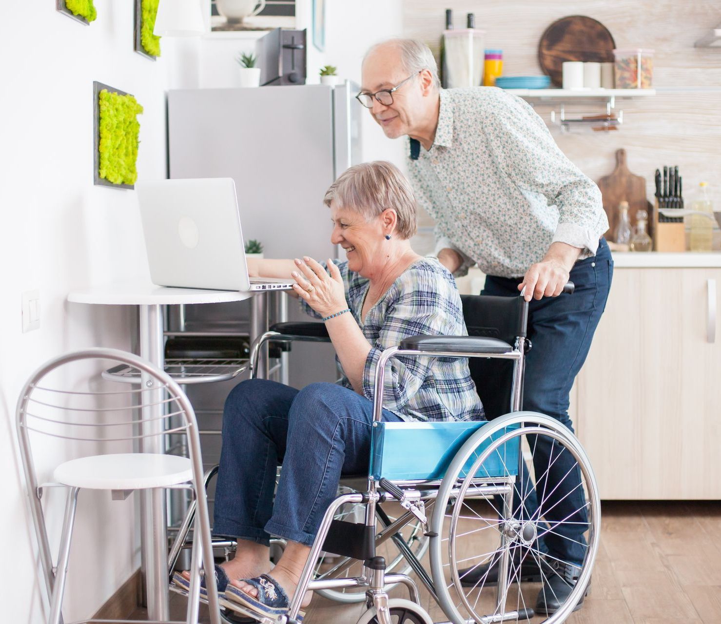 Older adult in a wheelchair using a laptop while a man stands beside her in a bright home office