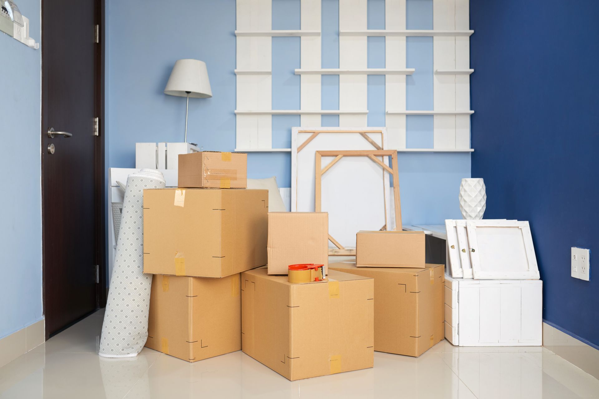 Stacked moving boxes in a blue room with shelves, a lamp, and a chair frame.