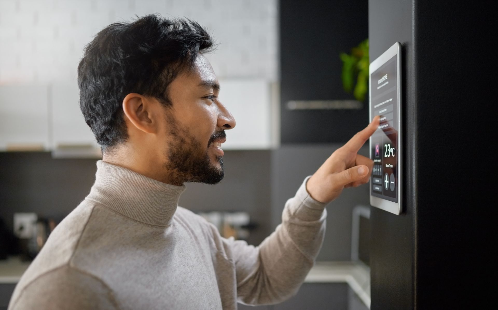 Person in a gray sweater taps a wall-mounted smart home display in a modern kitchen.