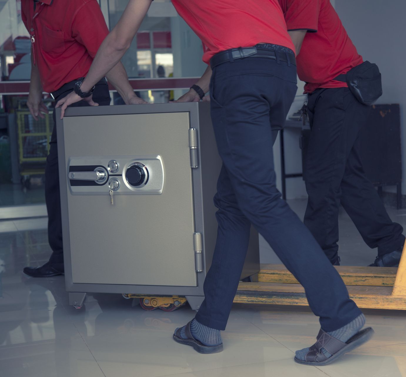 Workers in red shirts moving a large safe on a dolly through a building entrance