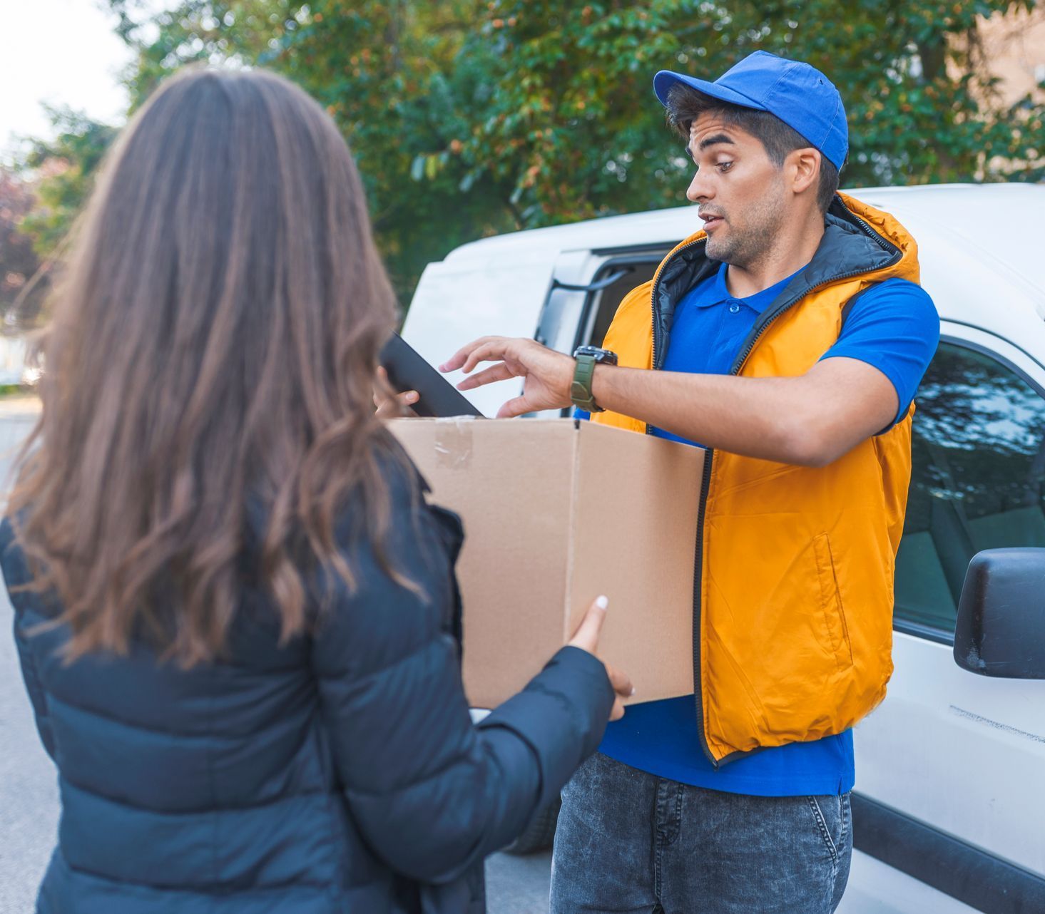 Delivery driver handing a package to a woman beside a white car outdoors