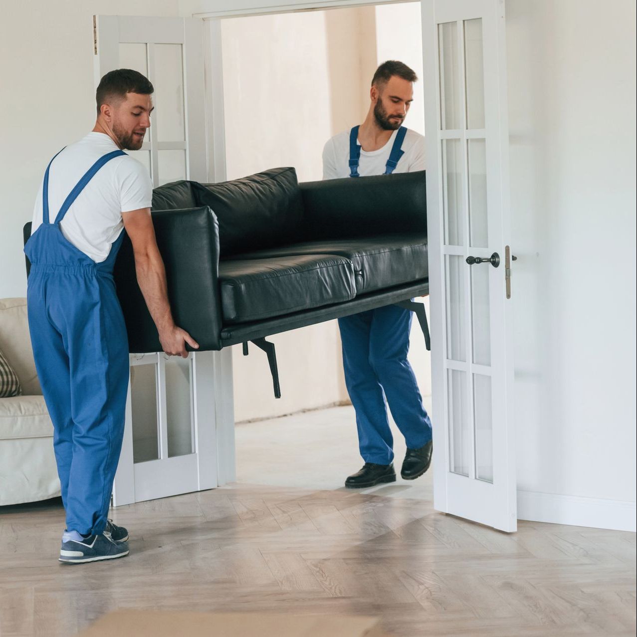 Two movers carrying a black sofa through a white doorway in a bright room