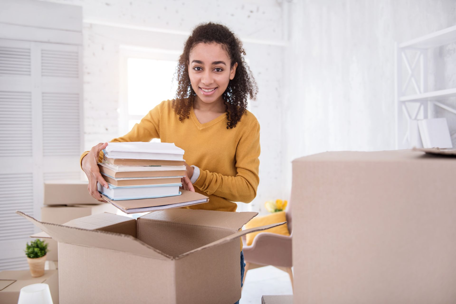 Woman packing books into a cardboard box in a bright room with moving boxes nearby