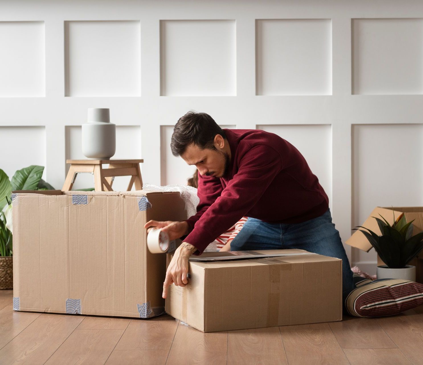 Man packing cardboard boxes in a bright room, kneeling beside a large moving box.