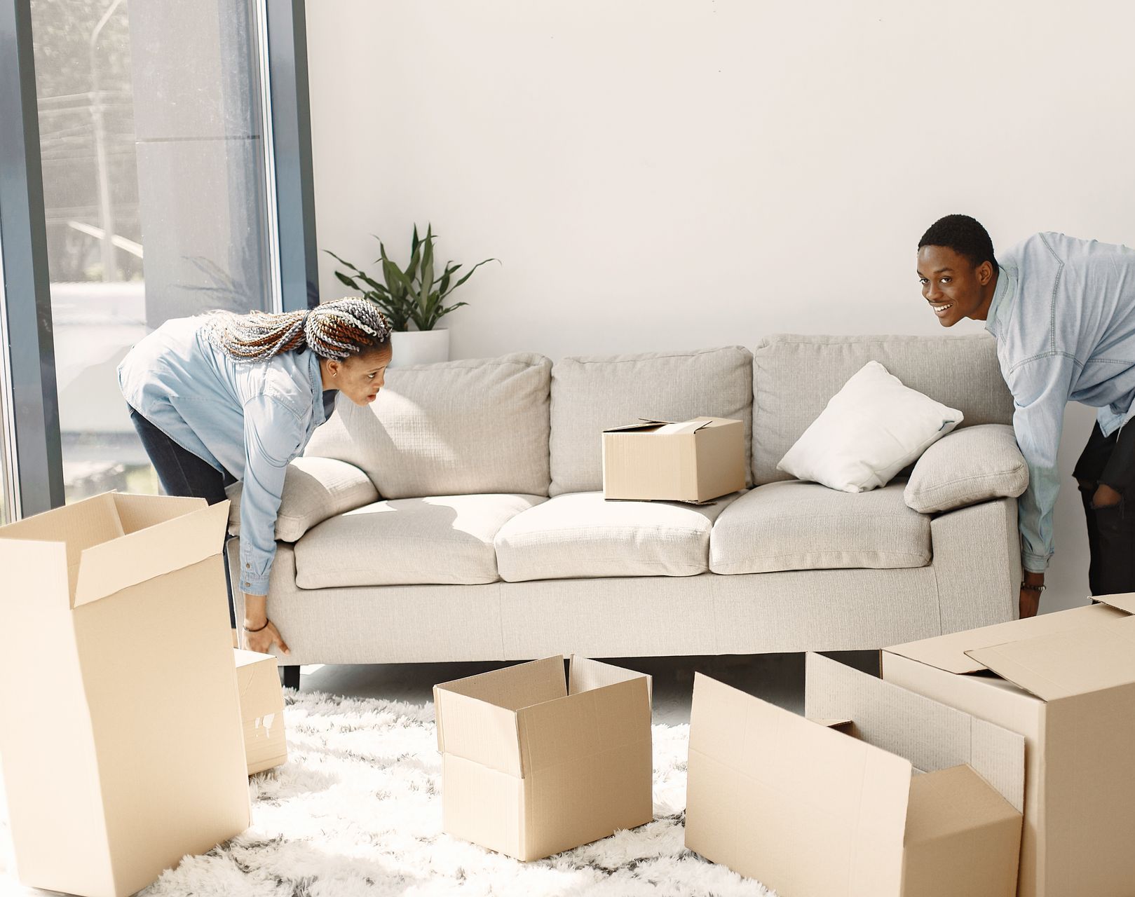 Two people moving a beige sofa into a bright living room with several cardboard boxes.