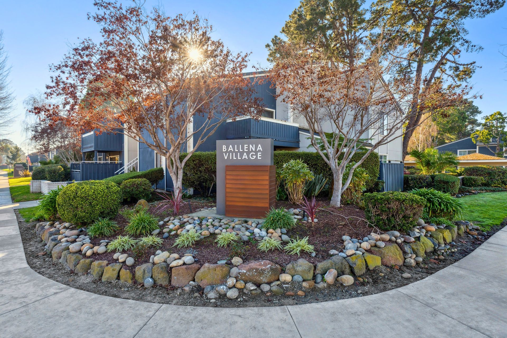 Modern residential building entrance with stone landscaping and a wooden sign at sunset at Ballena Village apartments in Alameda, CA.