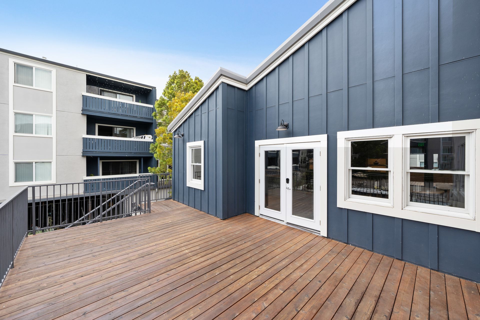 Wooden rooftop terrace beside blue-gray apartments with white-trimmed windows and a railing overlooking the courtyard at Ballena Village apartments in Alameda, CA.