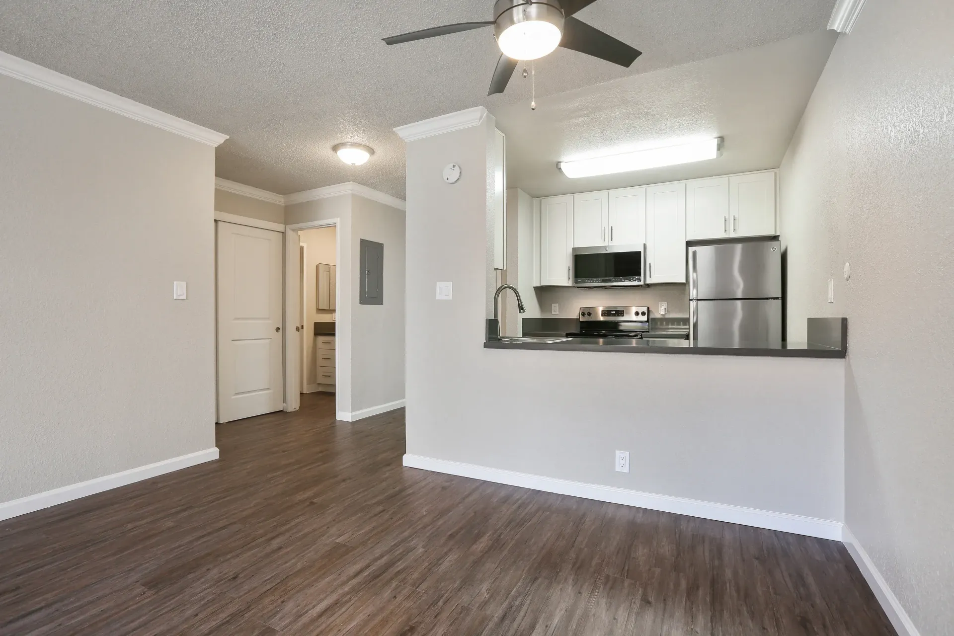 Empty living room with dark wood floors, ceiling fan, and open kitchen with stainless steel appliances at Ballena Village apartments in Alameda, CA.