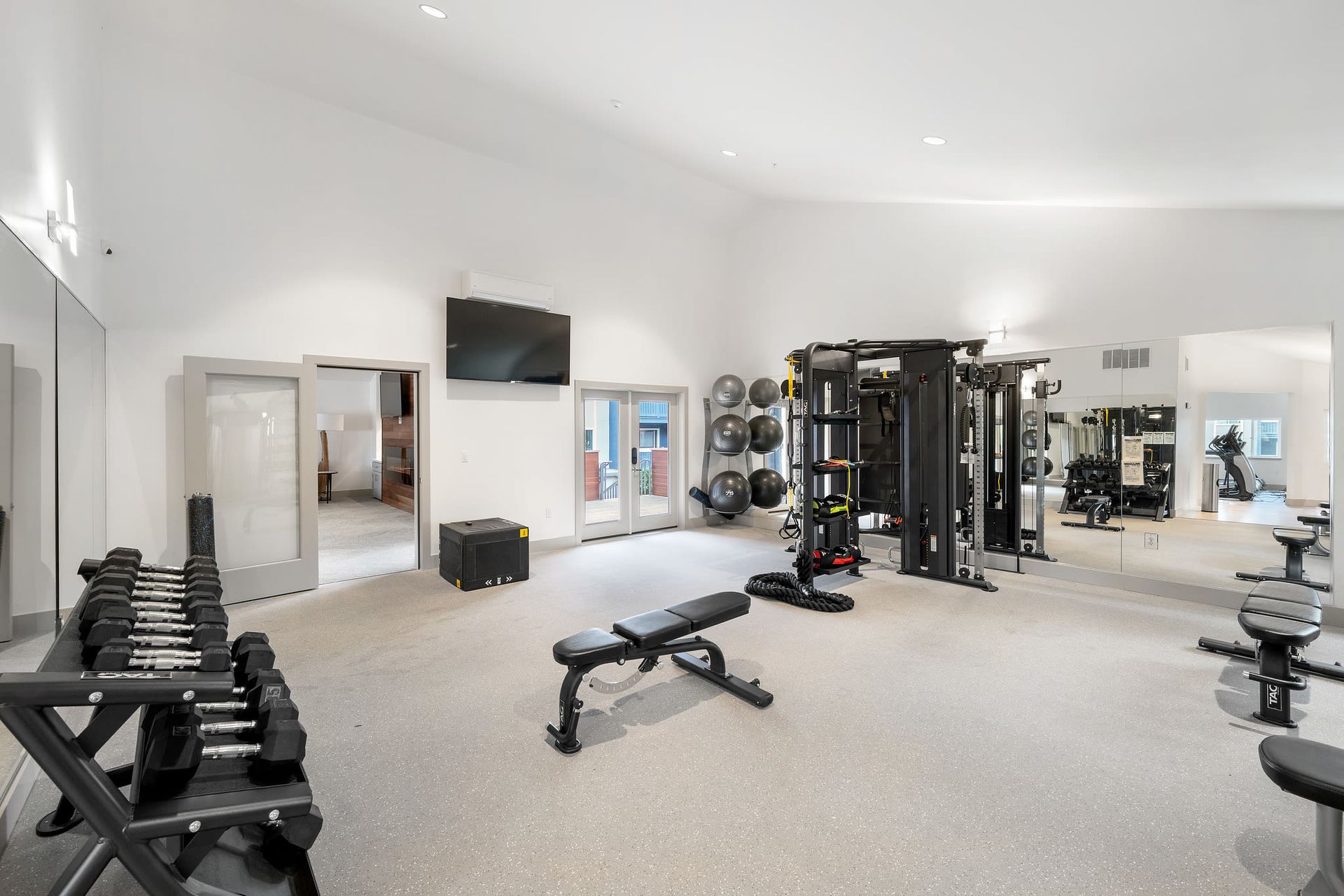Bright on-site community gym with free weights, benches, and a large mirrored wall at Ballena Village apartments in Alameda, CA.