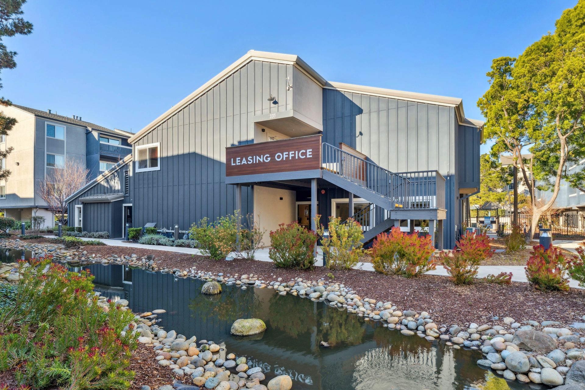 Leasing office building beside a small pond, with stone landscaping and blue sky at Ballena Village apartments in Alameda, CA.