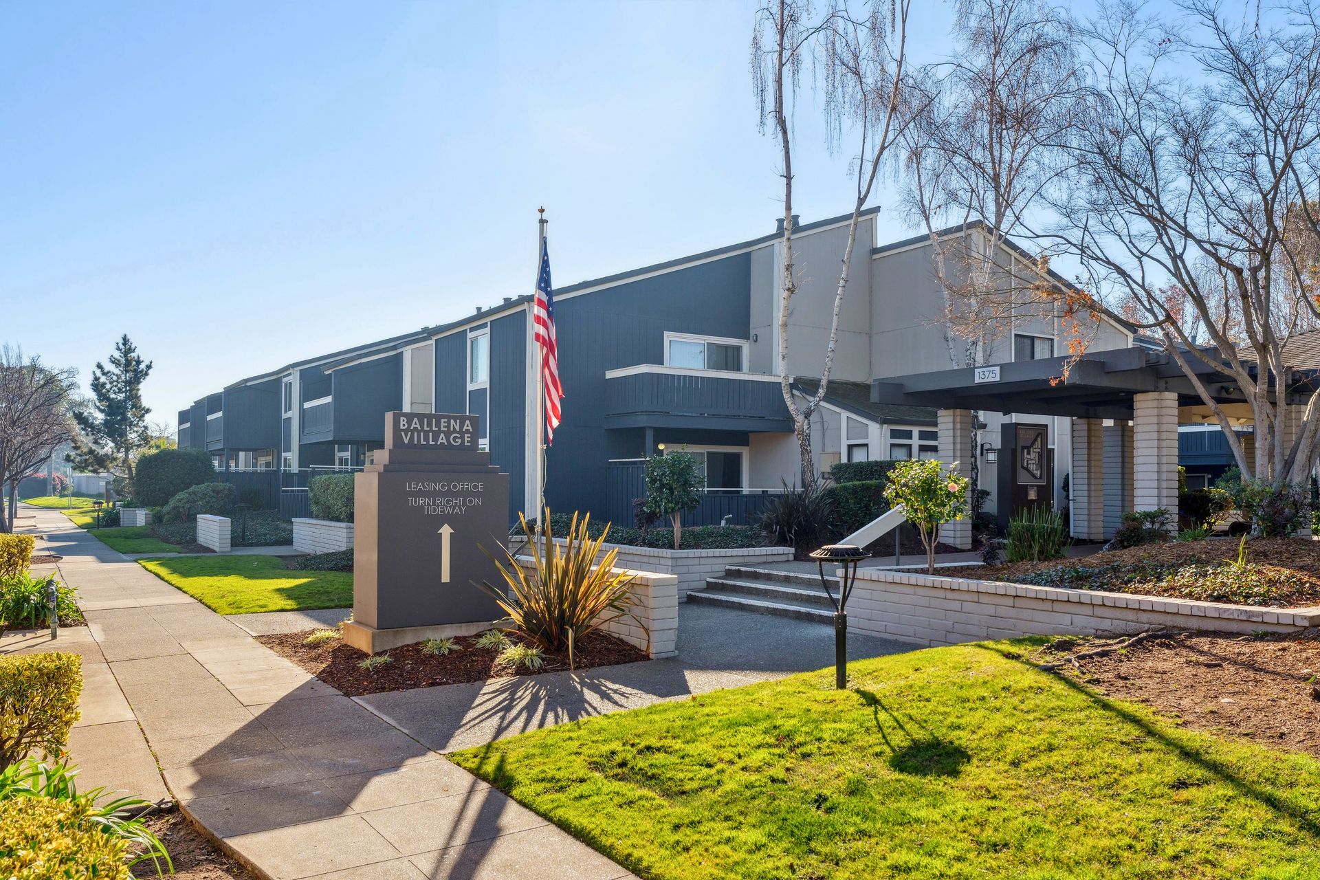 Modern building with a sign and American flag beside a sunlit sidewalk and landscaped entrance at Ballena Village apartments in Alameda, CA.