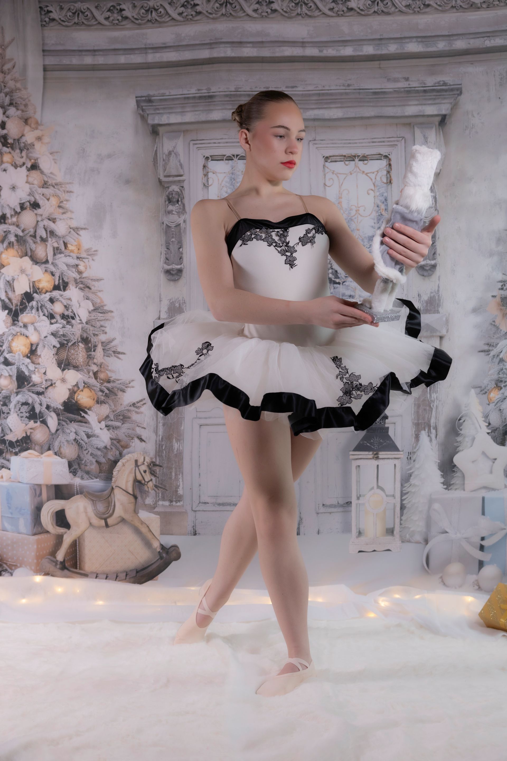 Ballerina in white tutu holds a nutcracker. Posed in snowy scene with Christmas tree and gifts.