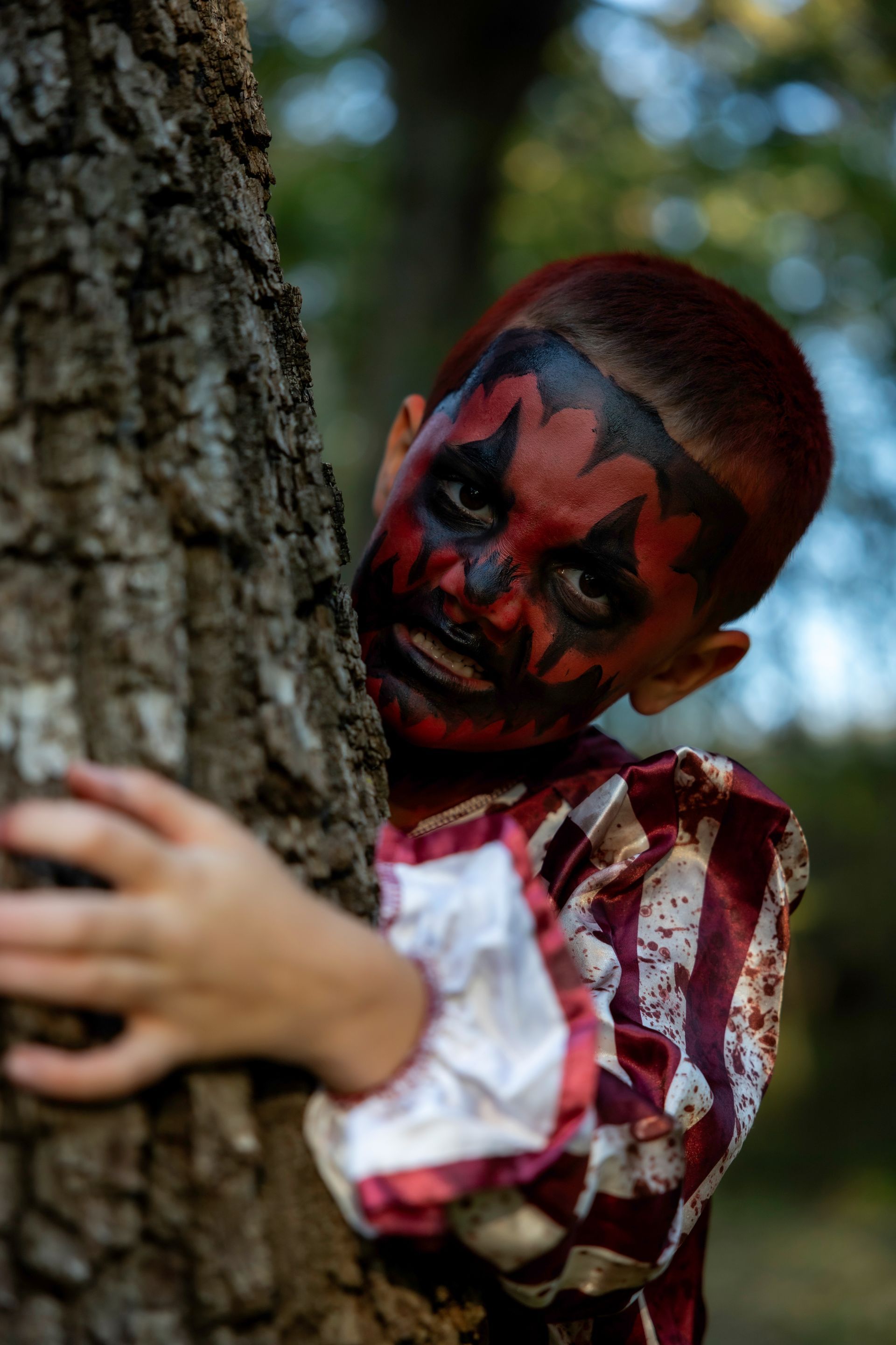 A young boy with his face painted like a clown is hugging a tree.